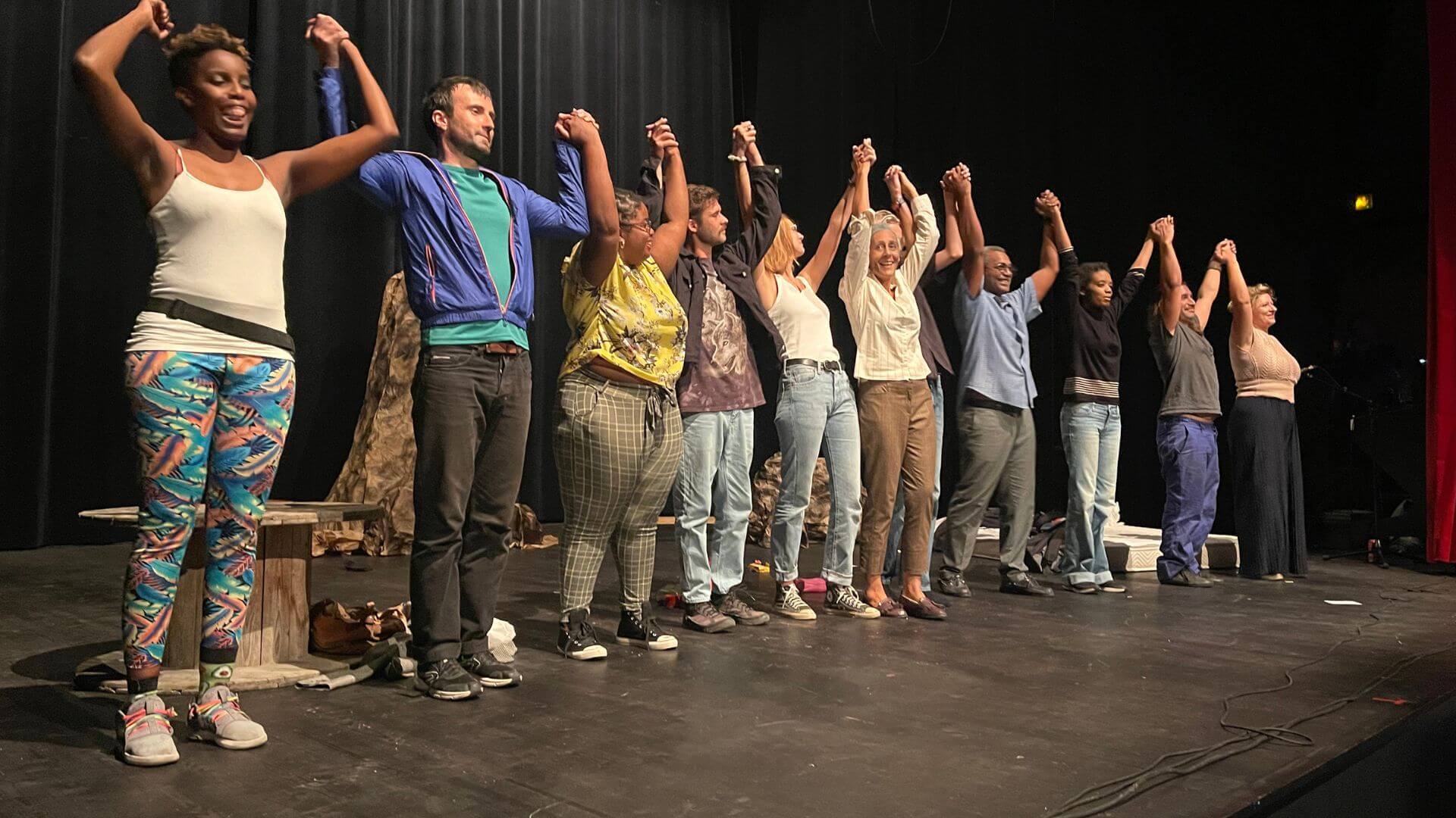 Groupe dynamique d'acteurs en formation, main dans la main, lors d'un exercice de cohésion à la fin d'un cours de théâtre en Guadeloupe. Ils affichent divers styles vestimentaires et expriment le plaisir et l'unité, reflétant l'esprit inclusif et la diversité culturelle de l'île. L'atmosphère est détendue et joyeuse, caractéristique des ateliers interactifs offerts dans la région.